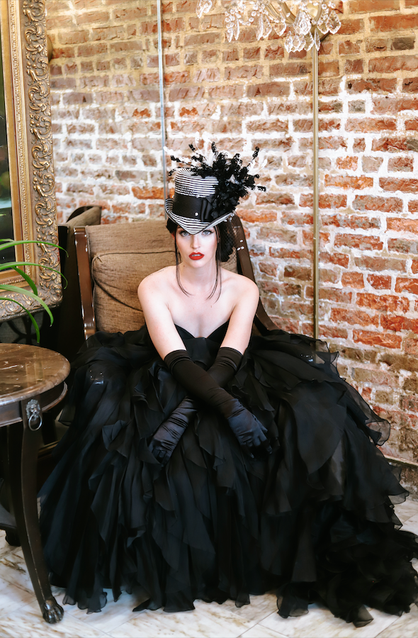 Woman in a black dress and hat sitting against a brick wall — Voodoo Makeup (Katy/Houston, TX)